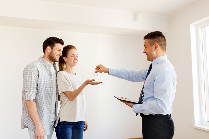 A man in a suit hands keys to a smiling couple inside a bright, empty room, possibly signifying the purchase or rental of a new home. The woman reaches out to accept the keys.