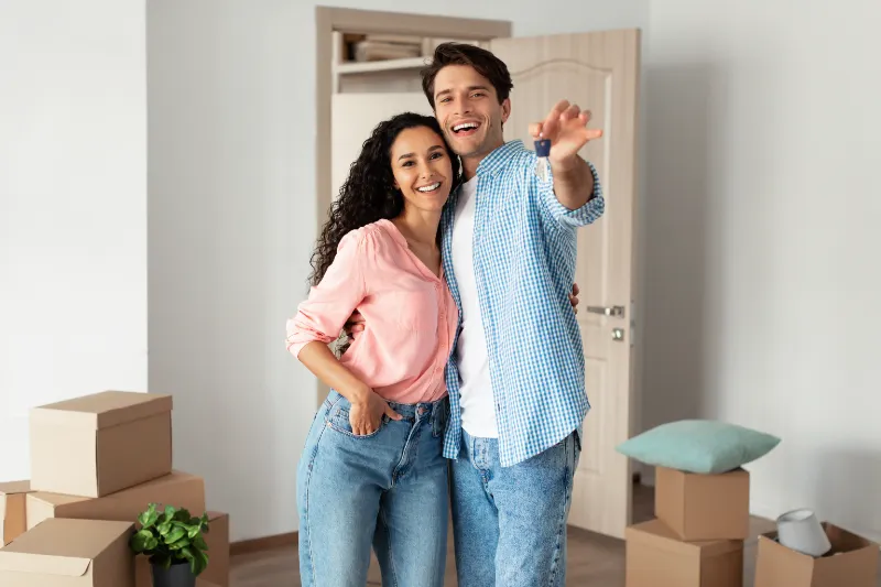 A smiling couple stands close together in a room with moving boxes. The woman wears a pink shirt and jeans, and the man wears a blue checkered shirt and jeans, gesturing excitedly as they look toward the camera.