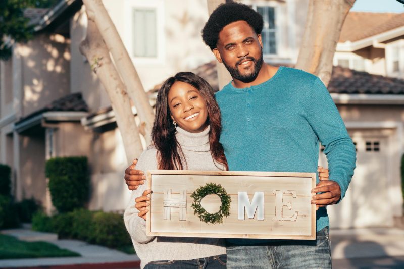 Happy couple holding a 'Home' sign in front of a charming new house, smiling together.