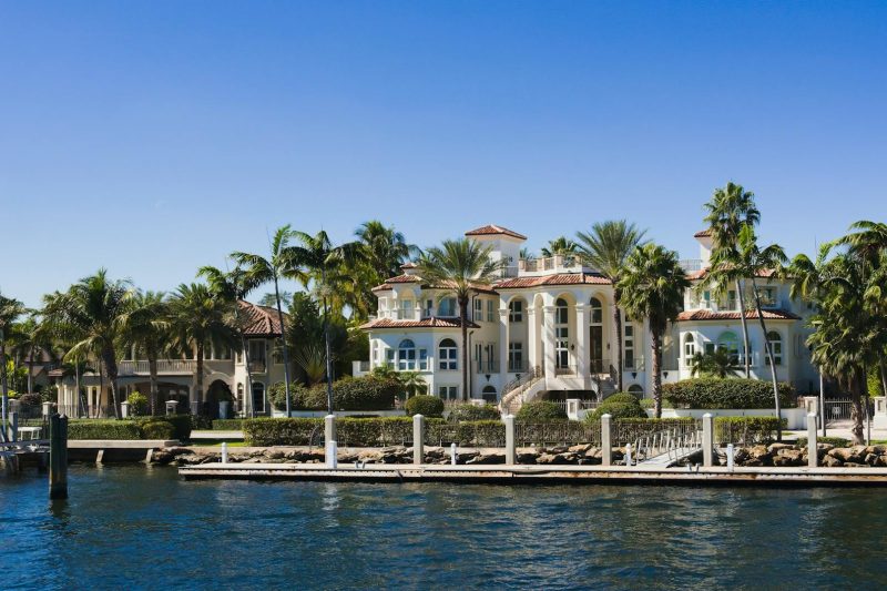 Elegant waterfront mansion with palm trees under a clear blue sky, exuding opulence and a coastal vibe.