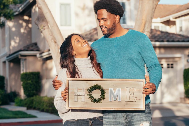 Joyful couple outdoors, holding a 'Home' sign, celebrating their new home purchase.