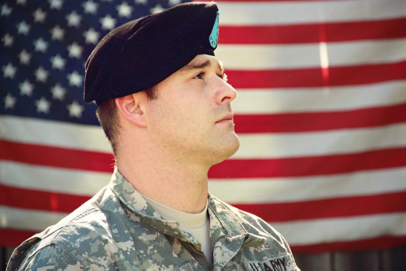 A US soldier in uniform stands proudly in front of the American flag, embodying patriotism.