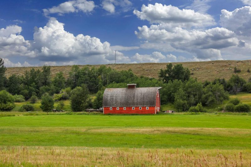 Red barn in lush green fields and hills under a cloudy blue sky in rural Montana.