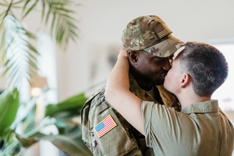 A loving military couple shares a kiss indoors, surrounded by warm lighting and a cozy home atmosphere.