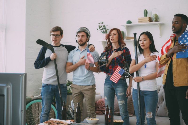 Home 15 Diverse friends watching sports on a TV while surrounded by American flags indoors.